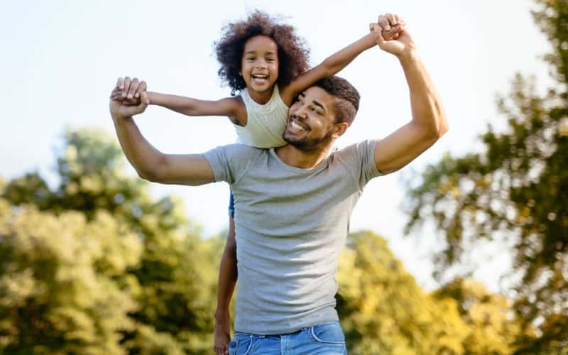 Black father holds his smiling daughter on his shoulders at the park